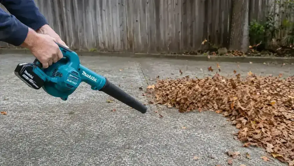 Close-up of a Makita DUB185Z cordless leaf blower being held at a low angle to sweep leaves across a concrete driveway